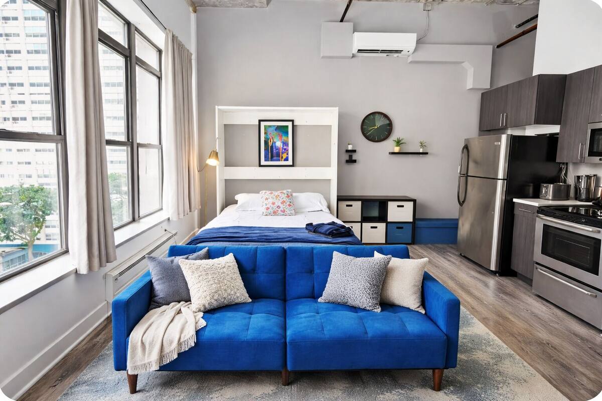 A modern apartment with a blue sofa, a bed, a kitchen, and a large window.