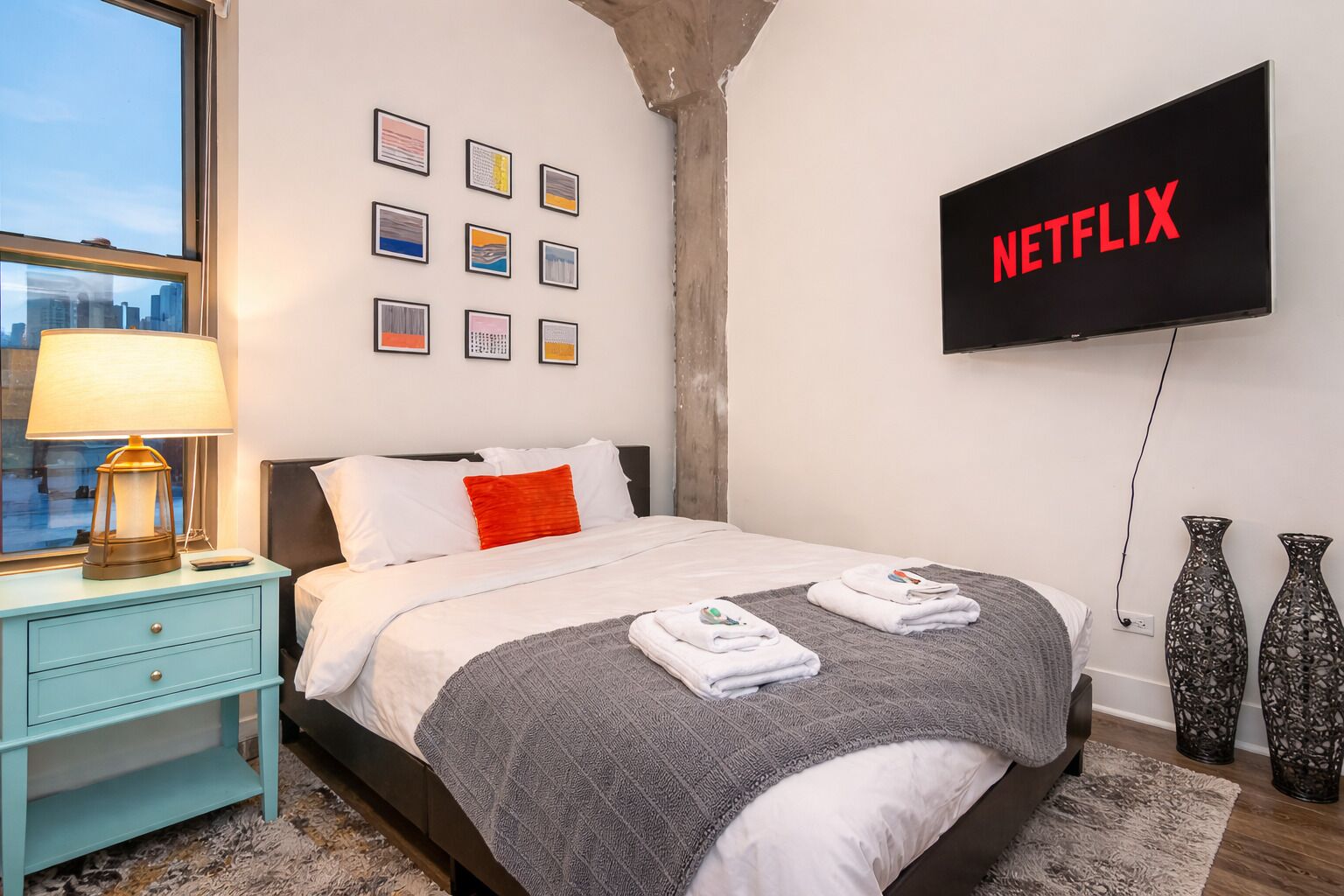 A hotel room with a bed, a TV mounted on the wall, a nightstand with a lamp, and a window with a view of the city.
