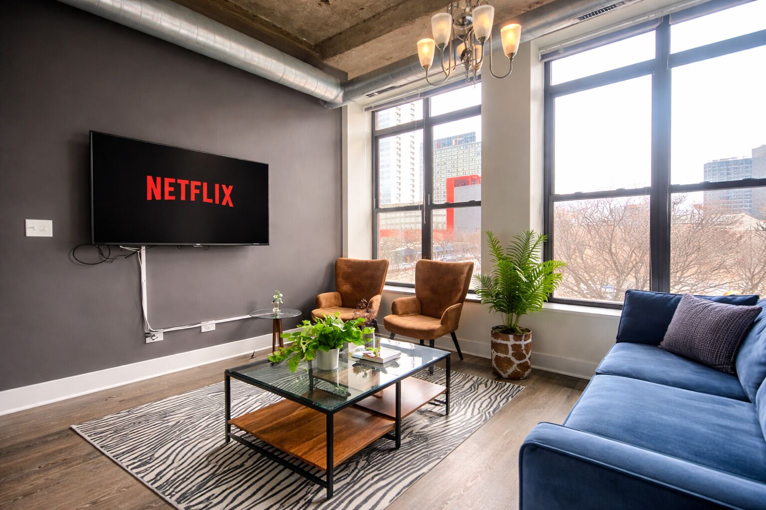 Un salon moderne doté d'un grand téléviseur à écran plat affichant le logo « NETFLIX », d'une table basse en verre, de deux fauteuils marron, d'un canapé bleu et d'une grande fenêtre offrant une vue sur la ville.