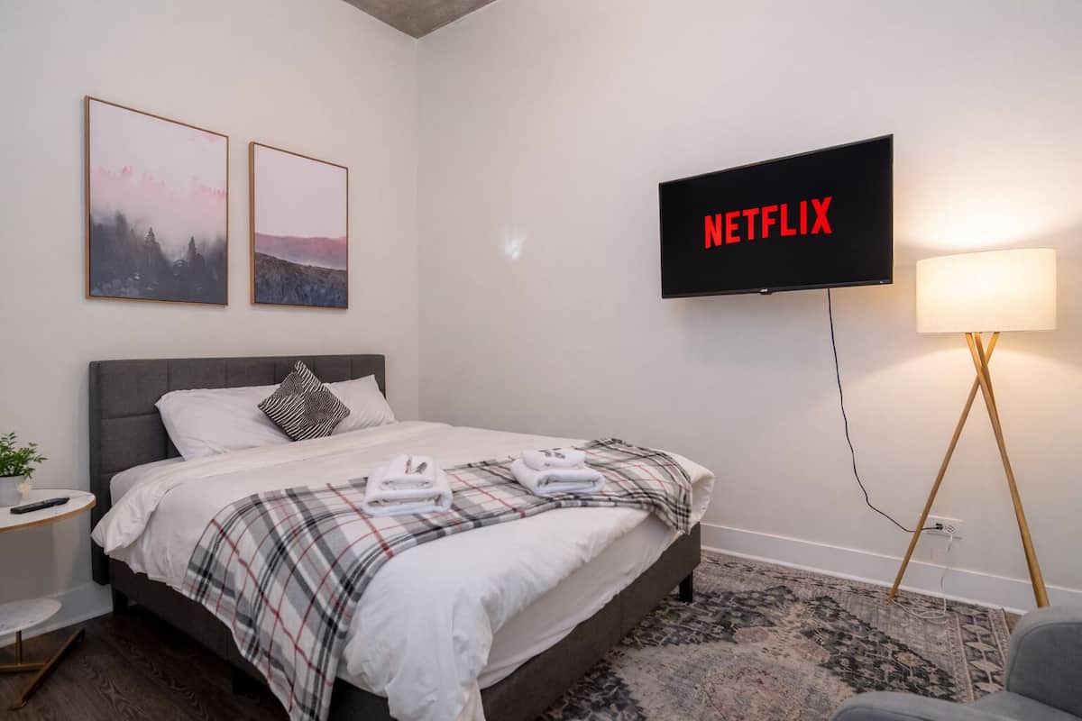 A hotel room with a bed, a TV mounted on the wall, a bedside table, and a lamp.