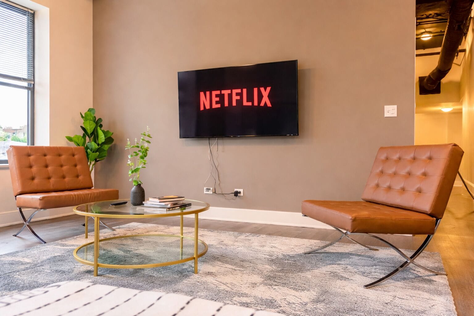 A modern living room with a television mounted on the wall displaying 'NETFLIX,' two leather armchairs, a glass coffee table, and a potted plant.