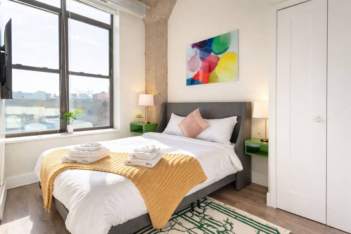 A hotel room with a bed, a window, a painting, and a green rug.