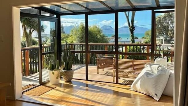 A room with a large window, wooden flooring, a bench, a potted plant, and a bed with pillows.