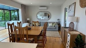 A dining area with a wooden table and chairs, a grey sofa, a round mirror, and a view of greenery outside.