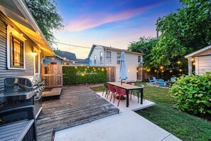 Un jardin avec une terrasse en bois, un barbecue, une table et des chaises d’extérieur, et des guirlandes lumineuses.