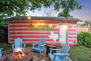 Una pequeña estructura de madera con un hoyo para fuego y sillas Adirondack azules, con la inscripción 'HOWDY HOUSTON, TX'.