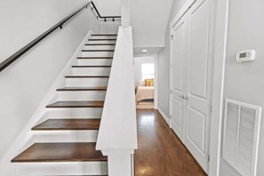 Un escalier avec des marches en bois et des murs blancs, menant à une pièce où se trouvent un lit et une fenêtre.