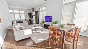 A modern living room with a sofa, dining table, and chairs, a kitchen in the background, and a TV mounted on the wall. - Houston Townhome w/ Skyline Views & Rooftop Patio (Houston)