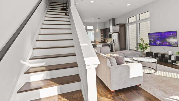 A modern living room with a staircase, a sofa, a TV, and a kitchen in the background. - Houston Townhome w/ Skyline Views & Rooftop Patio (Houston)