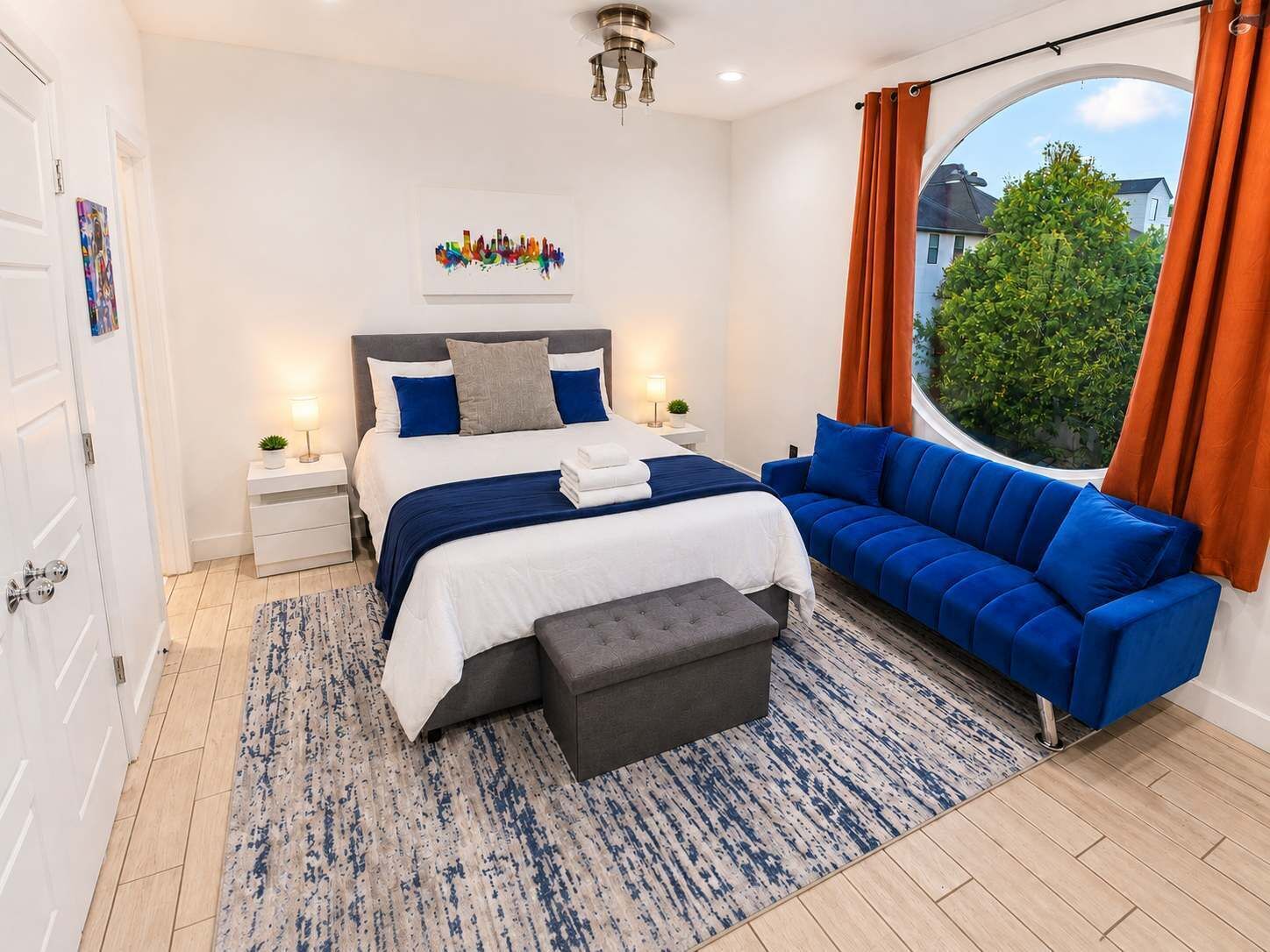 Quarto moderno com cama, sofá azul, banqueta cinza, mesa de cabeceira, abajur e janela com cortinas.