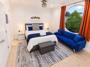 A modern bedroom with a bed, a blue sofa, a grey ottoman, a nightstand, a lamp, and a window with curtains. - Montrose Rooftop Townhome | 3 Suites, Sleeps 8 (Houston)