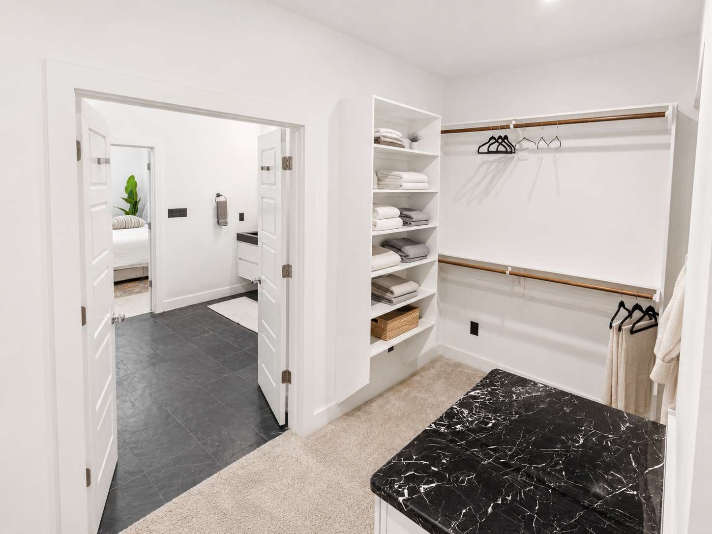 A modern bathroom with a black marble countertop, white storage shelves, and a walk-in closet with hanging clothes.