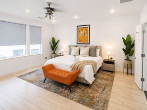 Ein Schlafzimmer mit Bett, Nachttisch, Stehlampe, einem Fenster mit Jalousien und Deckenventilator.
