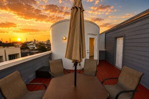 A rooftop terrace with a table and chairs, a water tank, and a view of the city skyline at sunset.