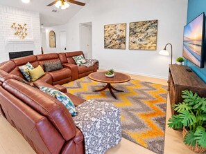 A living room with a brown leather sectional sofa, a round wooden coffee table, and a patterned ottoman. - 4 Queen Beds + Fire Pit | Energy Corridor (Houston)
