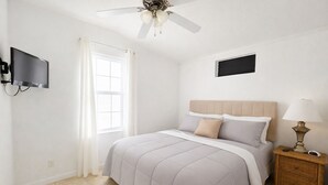 Une chambre à coucher avec un lit, une table de chevet avec une lampe, un ventilateur de plafond, un téléviseur fixé au mur et une fenêtre avec des rideaux.