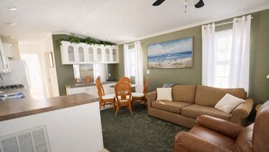 Un salon avec un canapé, une table à manger et des chaises, un coin cuisine et un tableau représentant une scène de plage.