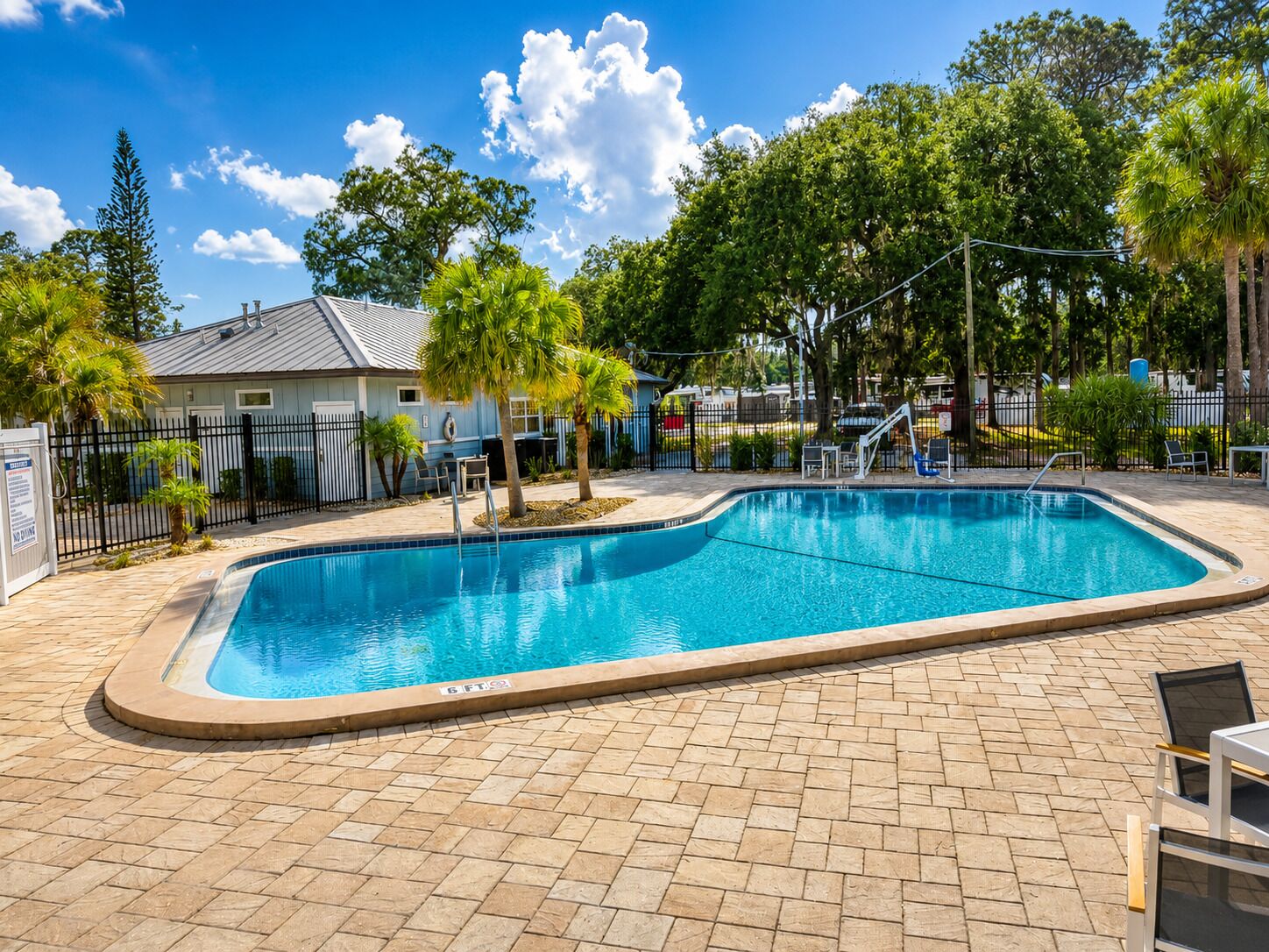 Une piscine rectangulaire, bordée d’une terrasse carrelée, entourée d’une clôture, d’arbres et d’un bâtiment en arrière-plan.