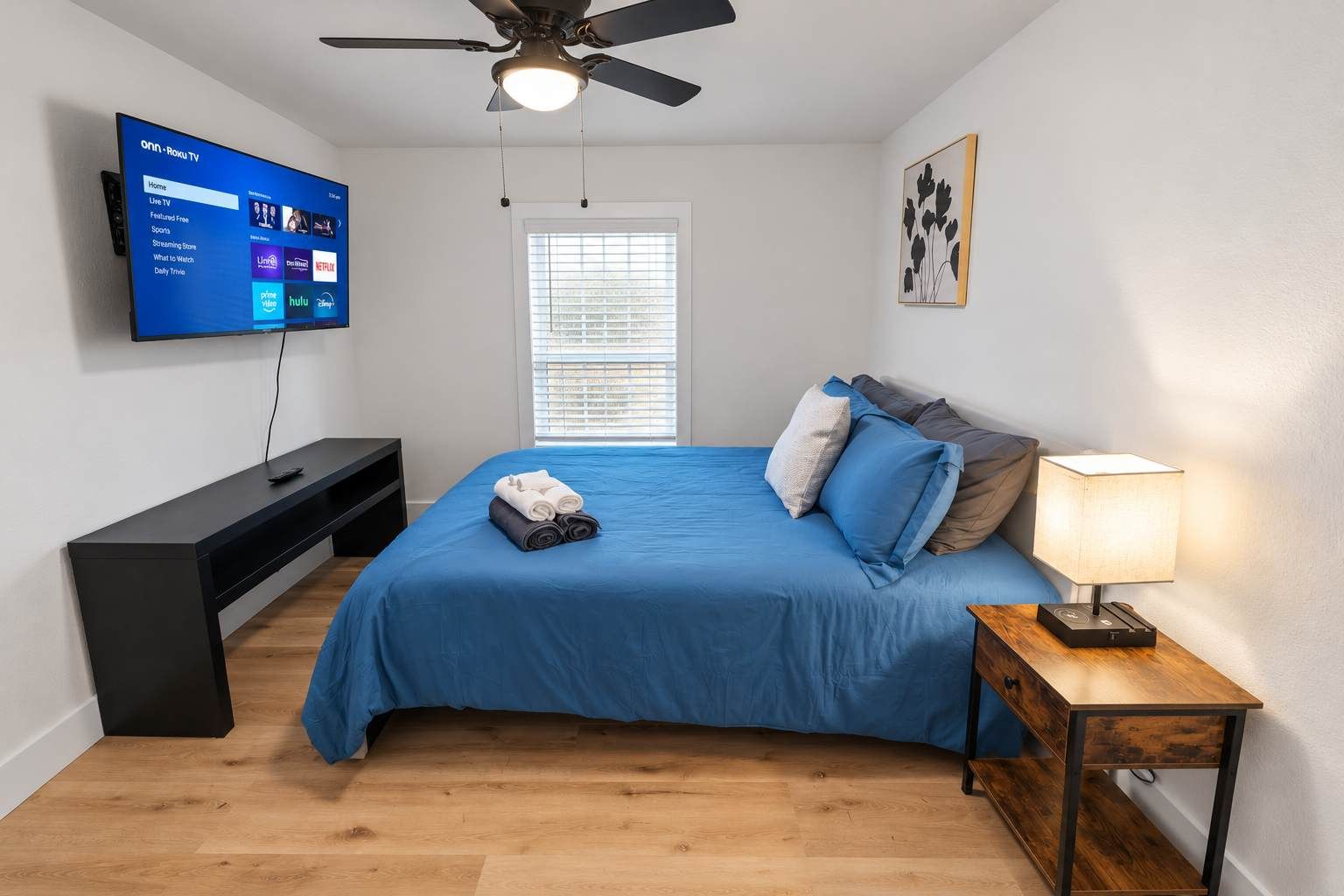 Un dormitorio con cama, televisión montada en la pared, ventilador de techo, mesita de noche con lámpara y ventana con persianas.