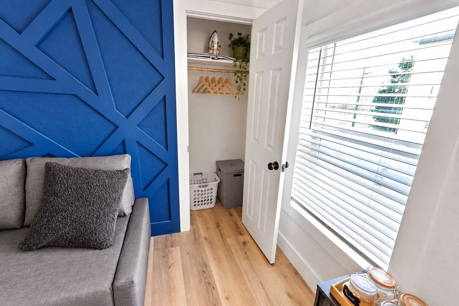 Une pièce avec un mur d’accent bleu, un canapé gris agrémenté d’un coussin texturé, un plancher en bois, une porte blanche avec une fenêtre et un panier à linge.