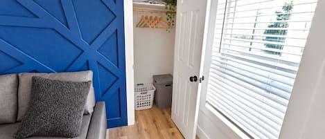 A room with a blue accent wall, a grey sofa with a textured pillow, a wooden floor, a white door with a window, and a laundry basket.