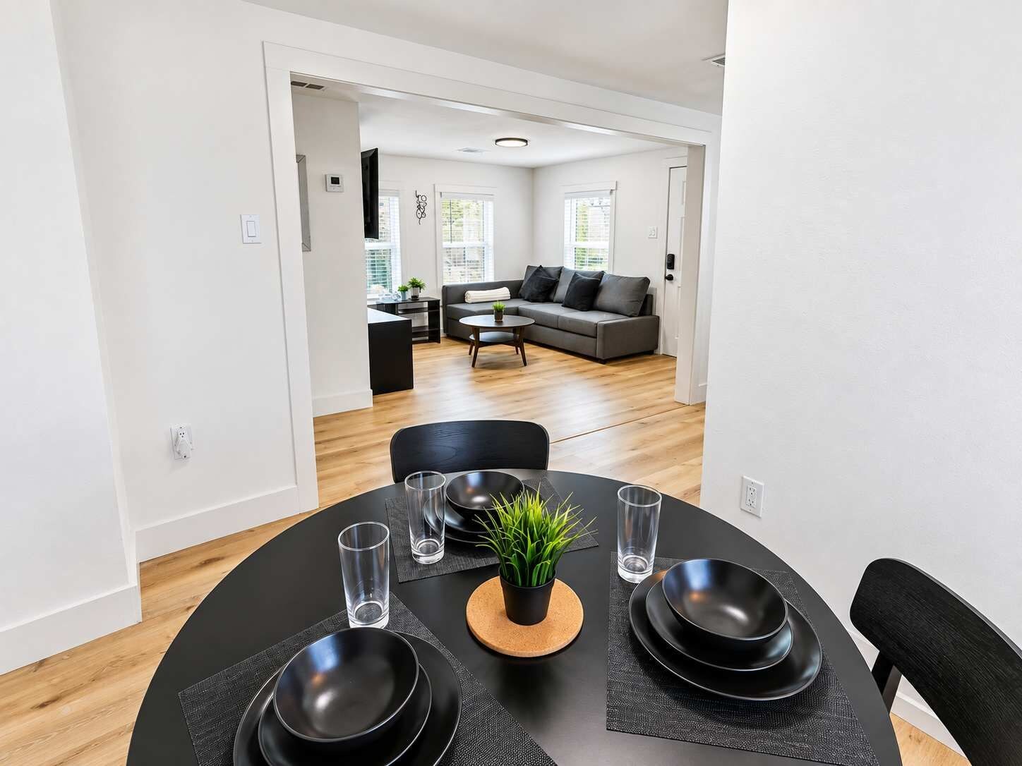 Un coin repas avec une table ronde, des assiettes noires et des verres, une petite plante en pot et, au fond, un canapé gris.