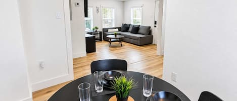 Ein Essbereich mit einem runden Tisch, schwarzen Tellern und Gläsern, einem kleinen Topfpflanzen und einem grauen Sofa im Hintergrund.