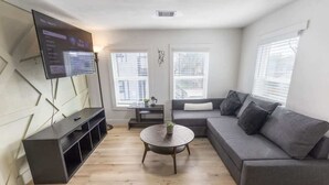 A modern living room with a grey sofa, a round coffee table, a flat-screen TV mounted on the wall, and a window with blinds. - 1BR Loft Near TSU + Med Center | Assigned Parking | Third Ward Houston 3 (Houston)