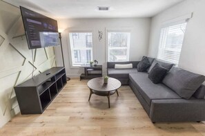 A modern living room with a grey sofa, a round coffee table, a flat-screen TV mounted on the wall, and a window with blinds. - 1BR Loft Near TSU + Med Center | Assigned Parking | Third Ward Houston 3 (Houston)