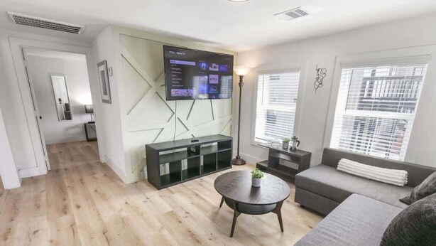 A modern living room with a flat-screen TV, a grey sofa, a round coffee table, and a bookshelf. - 1BR Loft Near TSU + Med Center | Assigned Parking | Third Ward Houston 3 (Houston)