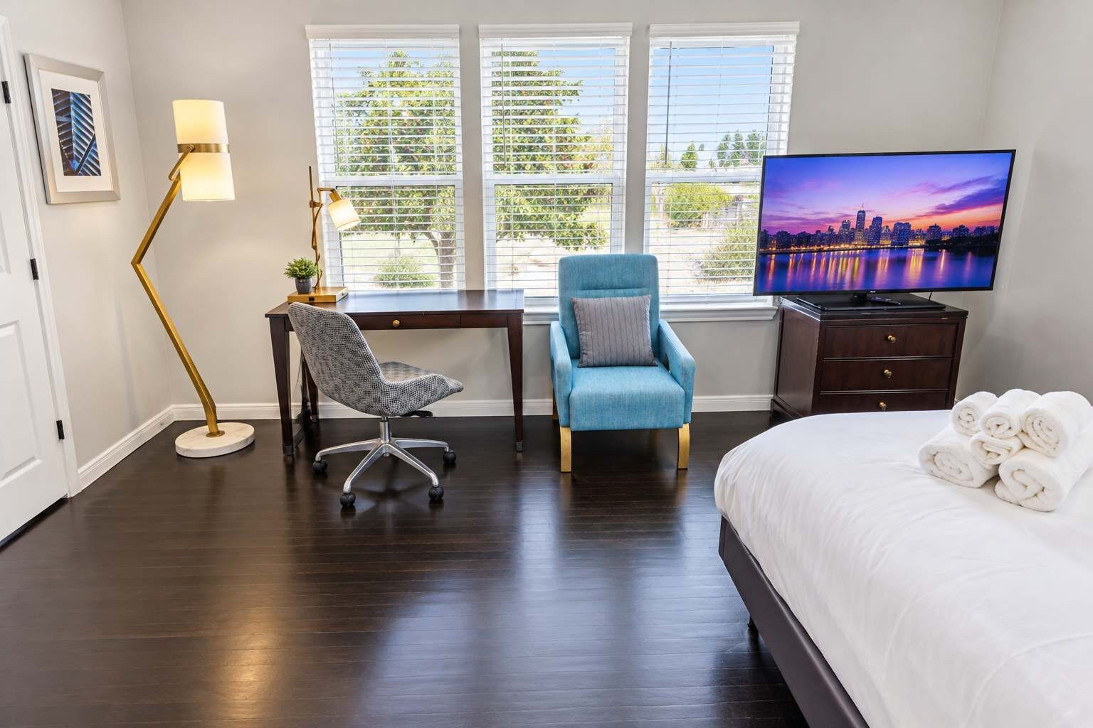 A hotel room with a bed, a desk, a chair, a TV, and a window with a cityscape view.