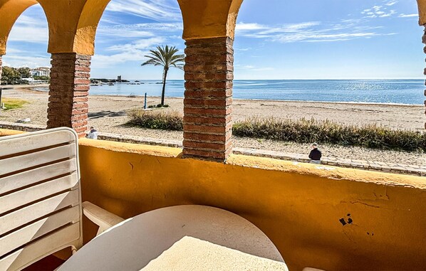 Vista desde un balcón con una mesa y una silla, con vista a una playa con una palmera y una persona sentada en la arena.