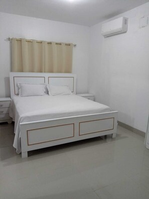 Uma cama branca com cabeceira, mesa de cabeceira e ar condicionado na parede.