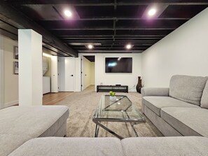 Ein modernes Wohnzimmer mit Glastisch, grauen Sofas, einem Flachbildfernseher und einem sichtbaren Waschbereich.