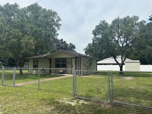 A single-story house with a front porch, a fenced yard, and a large tree in the background.