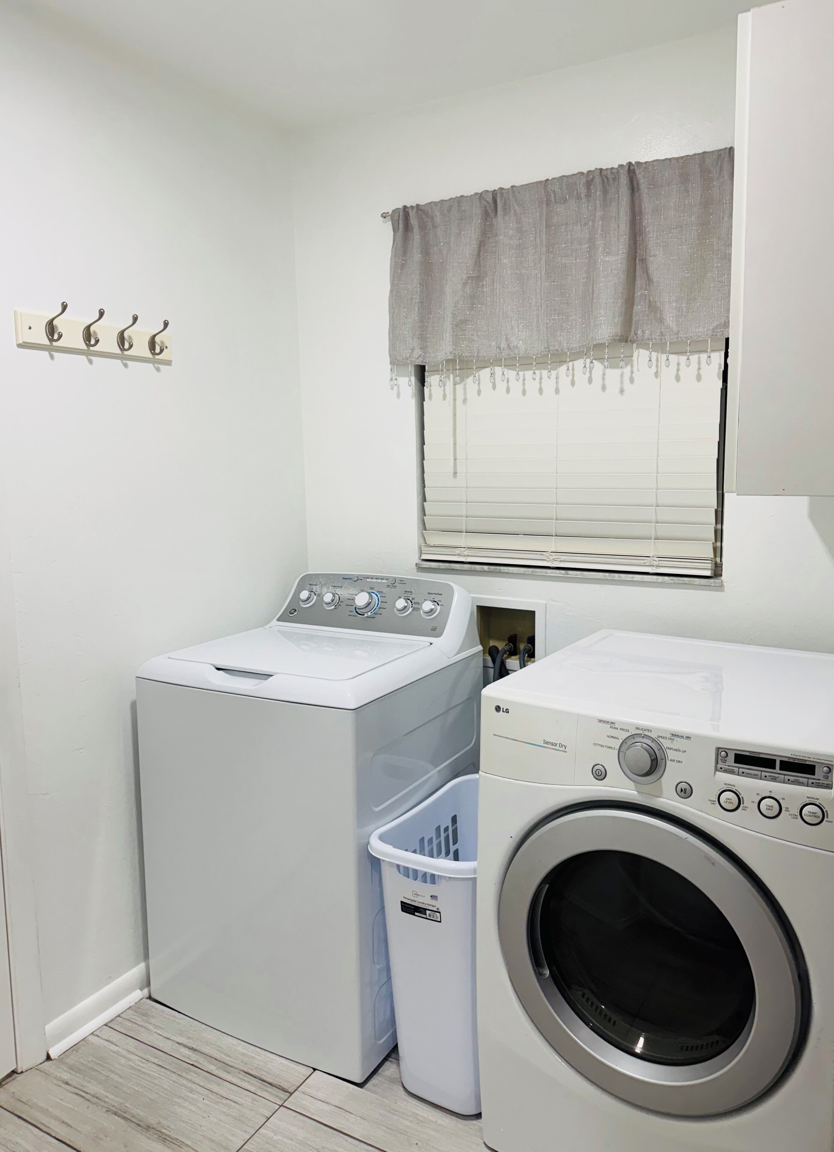 Een wasserette met een voorlader wasmachine en droger, een wasmand, een raam met gordijn, en een wandrek voor het drogen van was.