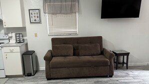 A living room with a brown sofa, a flat-screen TV, a kitchen area with a refrigerator, and a window with blinds. - Lumi House fully equipped house with everything you need for stay. (High Springs)