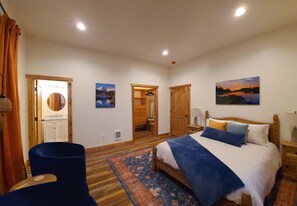 Ein Schlafzimmer mit einem Bett, einem Sessel, Holzboden, einem Gemälde und einer Tür zu einem Badezimmer.
