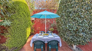 Une table dressée pour deux personnes, protégée par un parasol bleu, entourée de verdure et adossée à un mur rouge.