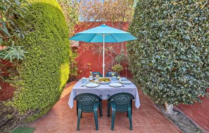 A table set for two with a blue umbrella, surrounded by greenery and a red wall.