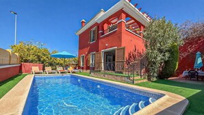 Une piscine avec un bord carrelé, un bâtiment rouge doté de balcons et une pelouse verte artificielle.