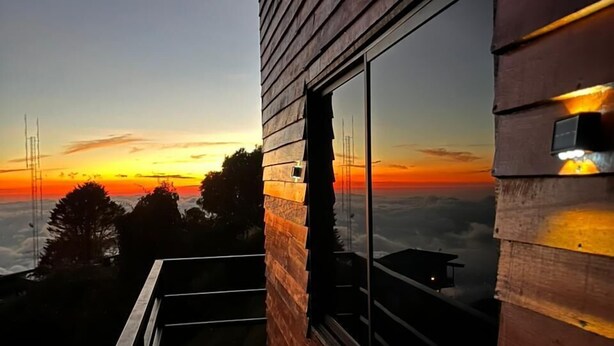 Fachada de madera con un amplio ventanal, un balcón y vistas al atardecer.