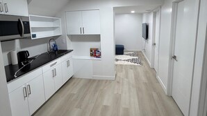 Et moderne køkken med hvide skabe, en sort bordplade og en mikrobølgeovn. Der er et lille siddeområde med et tæppe med mønster og et fjernsyn.
