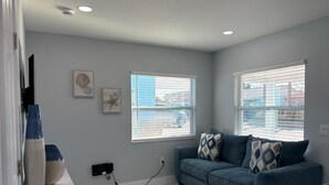 A room with a blue sofa, two windows with blinds, a wall-mounted air conditioner, and a power outlet. - Blue Tide Retreat, Walk to Beach, Perfect for Long Term Stays! (Madeira Beach)