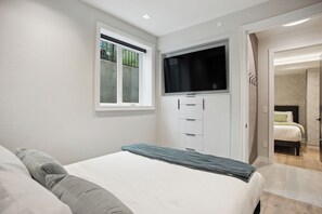Ein Schlafzimmer mit einem Bett, einem an einem Schrank montierten Fernseher, einem Fenster und einer Tür zu einem anderen Raum.