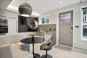 A modern kitchen with built-in appliances, a dining area with a round table and chairs, and a large window with blinds. - Luxury 2 Bedroom Downtown Beachside Unit (Vancouver)