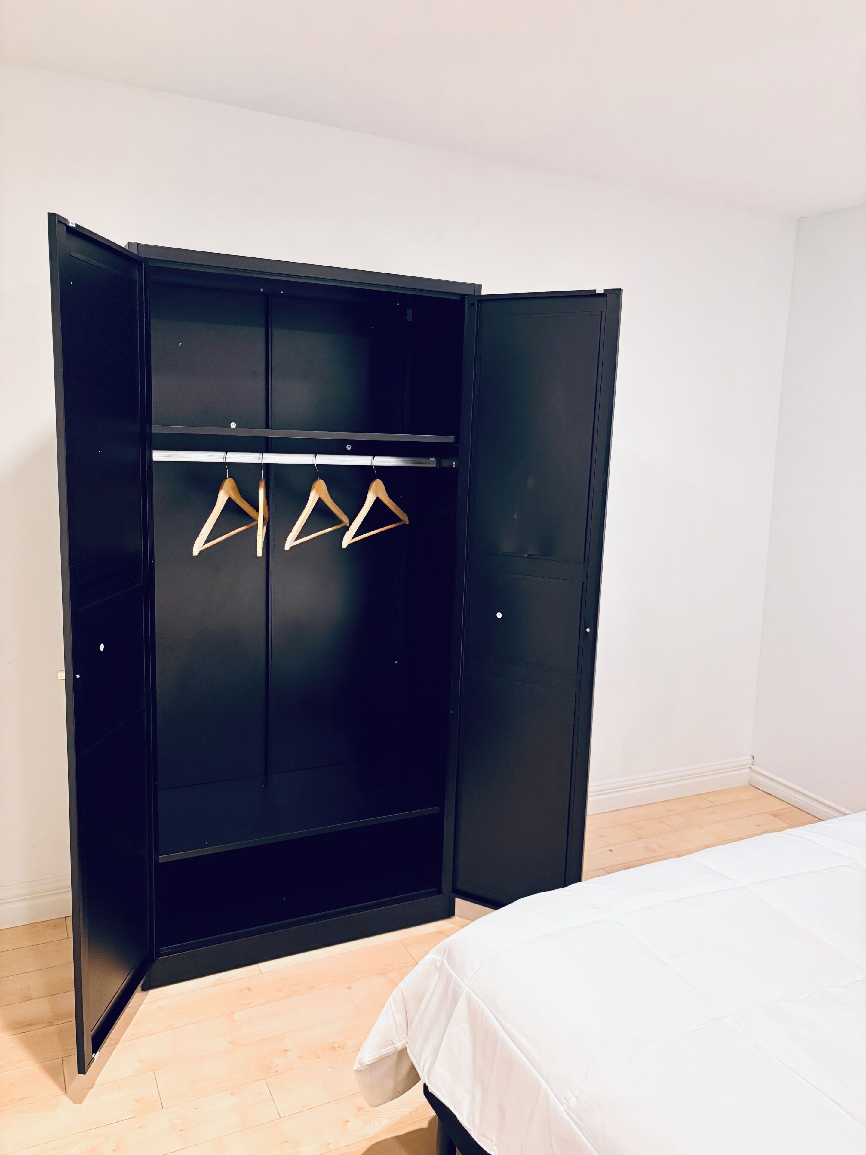 Une armoire noire avec trois cintres en bois, un lit avec une literie blanche et un plancher en bois.