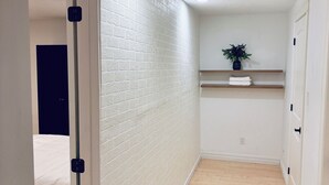 A narrow hallway with a white brick wall, wooden flooring, a ceiling light, a plant on a shelf, and a bed in the background. - Everything you need! spacious 1 BR |Full Kitchen |Bathtub,W/D wifi |Near Airport (Moncton)