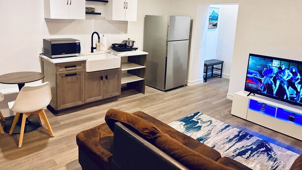 A modern kitchen with white cabinets, a microwave, and a refrigerator. There is a dining area with a table and chairs, and a living room with a sofa and a flat-screen TV. - Everything you need! spacious 1 BR |Full Kitchen |Bathtub,W/D wifi |Near Airport (Moncton)