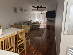 Ein Wohnzimmer mit einer Couch, einem Esstisch mit Stühlen, einem Deckenventilator und einem an der Wand befestigten Fernseher.
